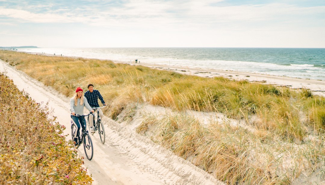 Dwie osoby jadą na rowerze wzdłuż piaszczystej ścieżki wydmowej na wybrzeżu Hiddensee, podczas gdy fale rozbijają się o plażę, a trawa kołysze się na wietrze. // Wycieczka rowerowa wzdłuż szerokich wydm odkrywa cichy, tętniący życiem nadmorski krajobraz Hiddensee i pozwala odetchnąć słonym wiatrem. // © MV-T/Petermann Dwie osoby jadą na rowerze wzdłuż piaszczystej ścieżki wydmowej na wybrzeżu Hiddensee, podczas gdy fale rozbijają się o plażę, a trawa kołysze się na wietrze.