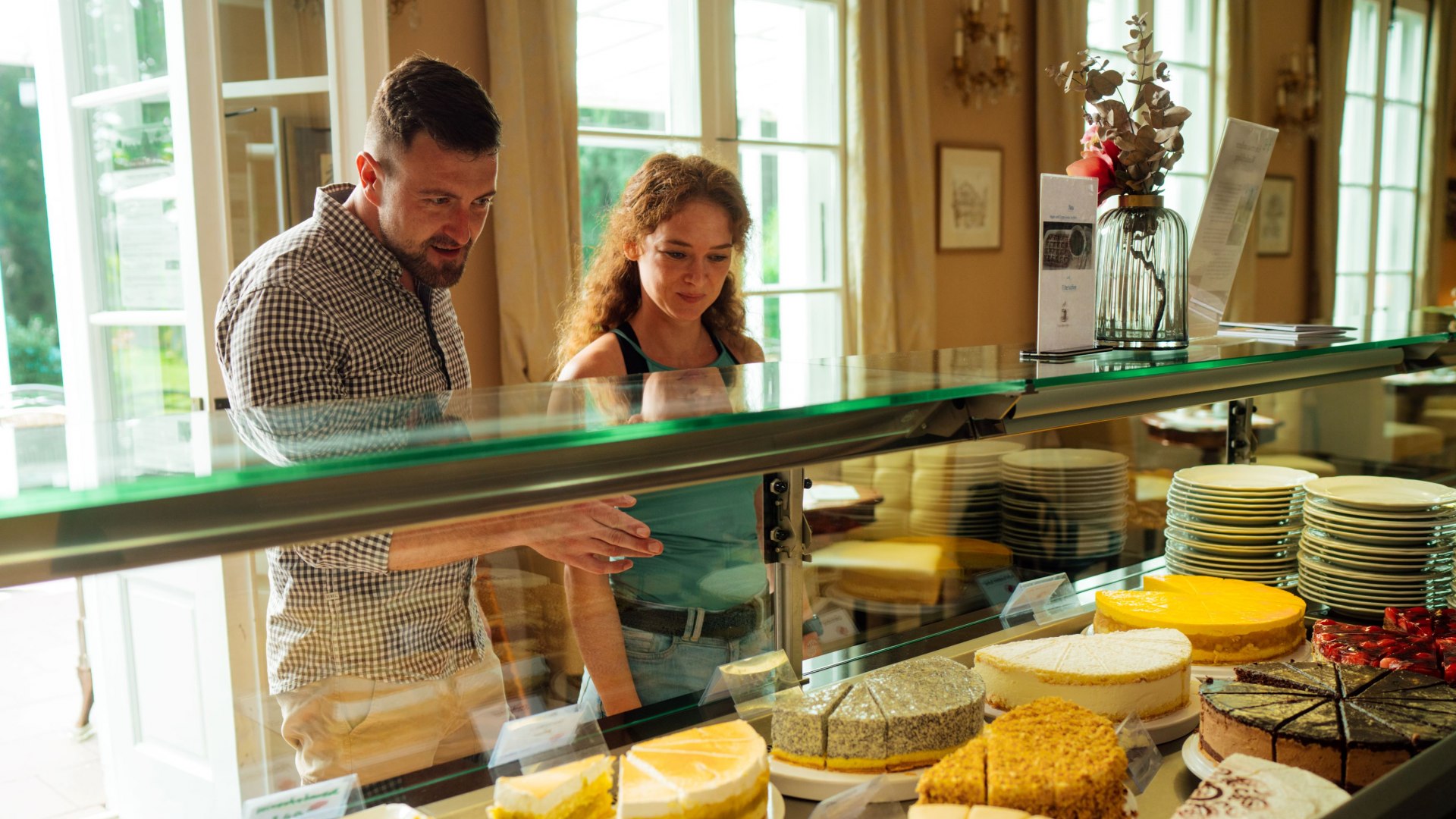A couple stands in front of a cake counter in the Rosencafé Putbus and looks at the selection of different cakes and pastries. The counter is richly stocked with freshly baked goods, including fruit tarts, cheesecakes and chocolate specialties. In the background, bright curtains and classic decorations create a cozy atmosphere.