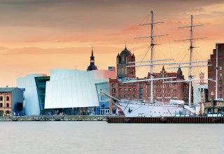 Modern museum architecture meets Hanseatic heritage: the Ozeaneum at Stralsund harbor merges with historic brick buildings and sailing ships to create a unique silhouette. In the evening light, the UNESCO World Heritage city glows in warm tones. Here you can experience maritime history up close - from the museum deck to the harbor promenade., &copy; Francesco Carovillano