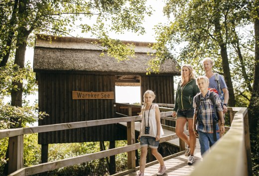 En familie går over en træbro hen til en hytte med stråtag; en pige har en kikkert hængende om halsen. // Knirkende træplanker, raslende løv: Ved Warnker-søen i Müritz-nationalparken udforsker familier gangbroen, der fører hen til hytten med stråtag. // © MV-T/Roth En familie går over en træbro hen til en hytte med stråtag; en pige har en kikkert hængende om halsen.