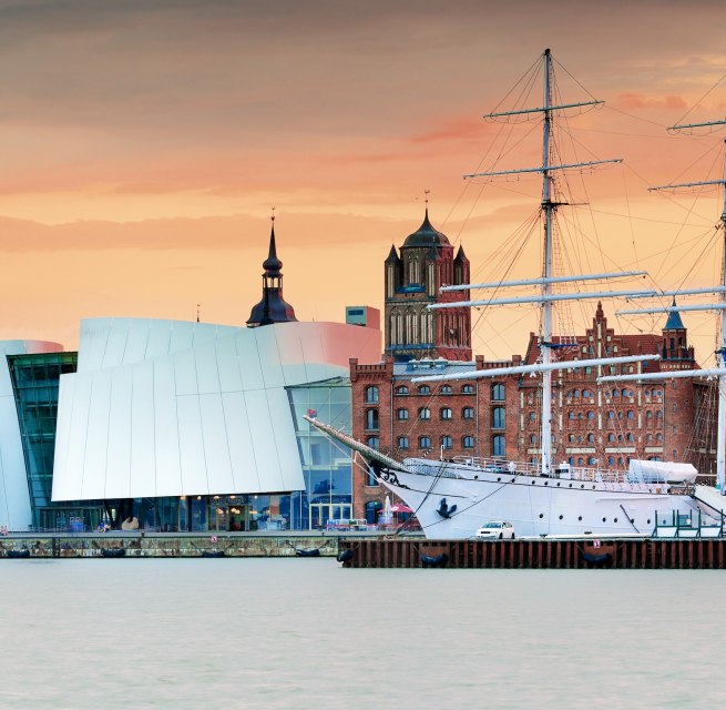 Modern museum architecture meets Hanseatic heritage: the Ozeaneum at Stralsund harbor merges with historic brick buildings and sailing ships to create a unique silhouette. In the evening light, the UNESCO World Heritage city glows in warm tones. Here you can experience maritime history up close - from the museum deck to the harbor promenade., &copy; Francesco Carovillano