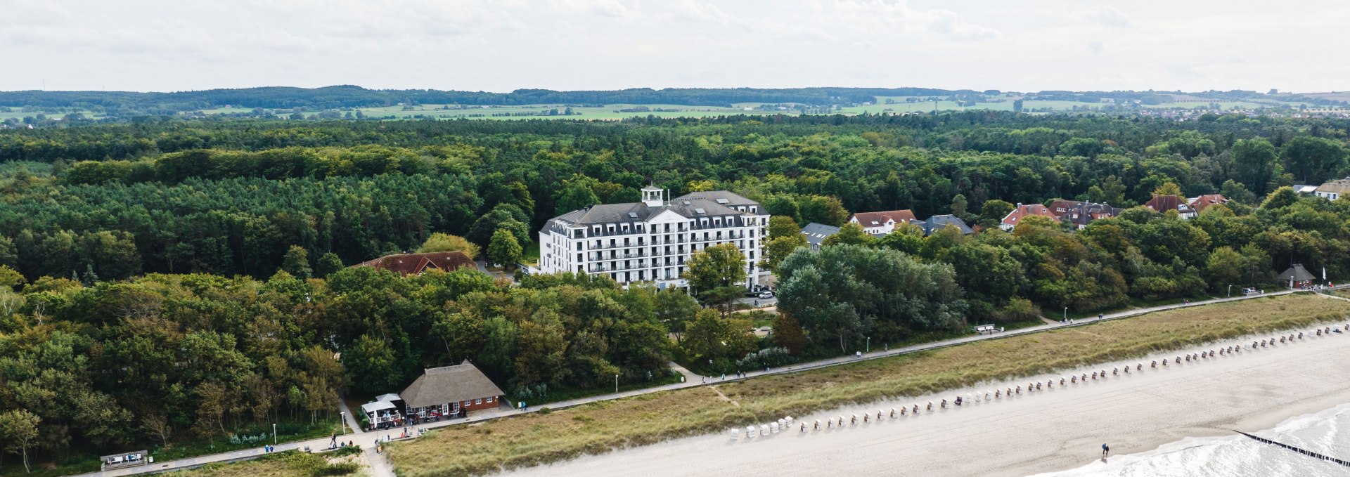 Widok z lotu ptaka na plażę i hotel przy plaży w K&uuml;hlungsborn, otoczony lasem, z leżakami i spokojną wodą Morza Bałtyckiego.