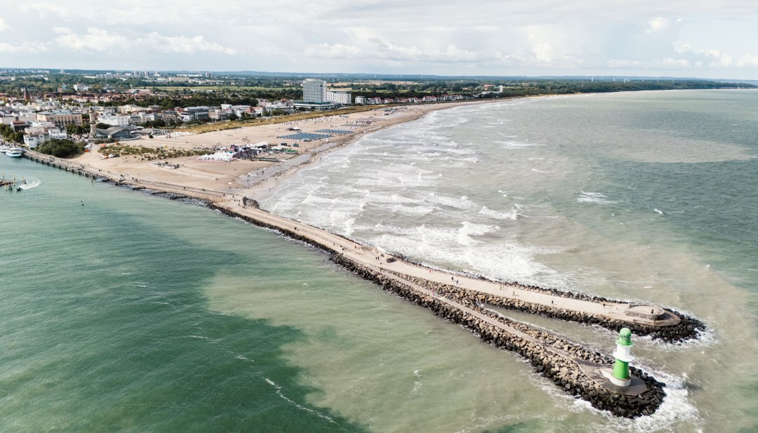 Widok z lotu ptaka na plażę Warnemünde z molo, latarnią morską i Morzem Bałtyckim w łagodnej fali. // Morze Bałtyckie obija się łagodnymi falami o szeroką piaszczystą plażę Warnemünde. Do morza prowadzi kamienne molo, zwieńczone zielono-białą latarnią morską. Wzdłuż wybrzeża ustawione są leżaki, a za nimi widać sylwetkę nadmorskiego kurortu z jego zabytkowymi budynkami i nowoczesnymi hotelami. // © MV-T/Scholz-Witzel Widok z lotu ptaka na plażę Warnemünde z molo, latarnią morską i Morzem Bałtyckim w łagodnej fali.