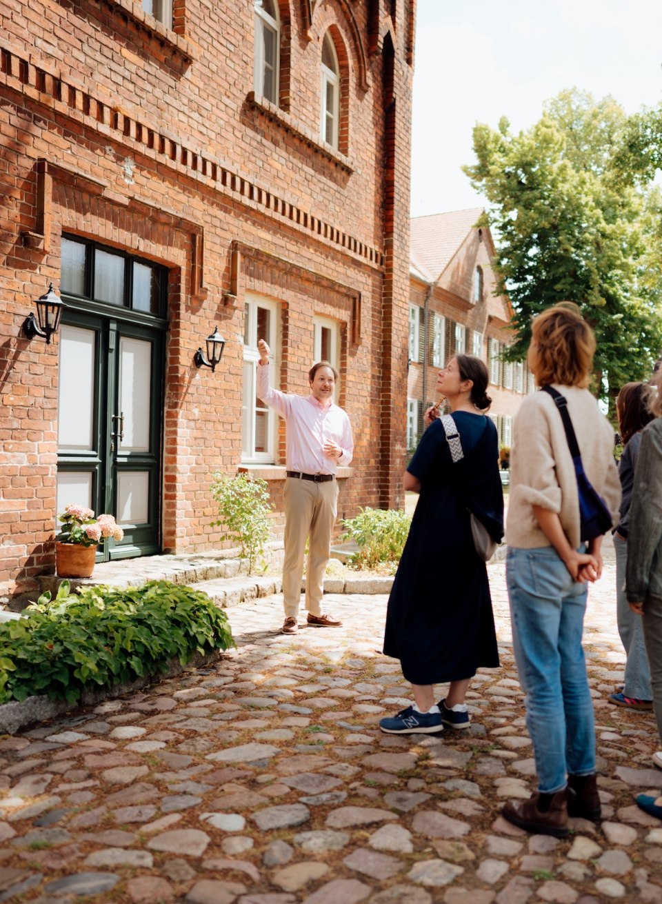 Jannis Achtenhagen explains the brick construction of the Ludorf manor house to a group on a cobblestone path surrounded by greenery. // Amid brick buildings and old trees, Jannis Achtenhagen leads the group around the Ludorf manor house, pointing out details on the facade and offering new perspectives // © MV-T/Petermann Jannis Achtenhagen explains the brick construction of the Ludorf manor house to a group on a cobblestone path surrounded by greenery.