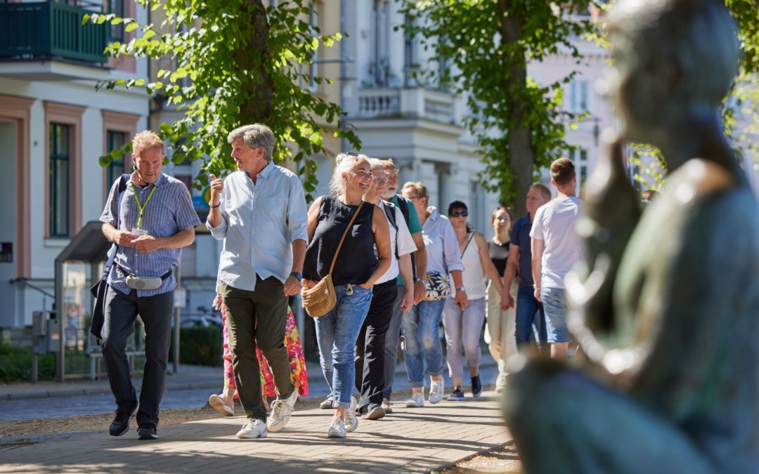 A group of people are walking along a cobbled path with a city guide. On the right, blurred in the picture, a statue can be seen by the Pfaffenteich pond; the villas appear in the background. The focus is on the guide and a couple having a lively conversation., © Oliver Borchert