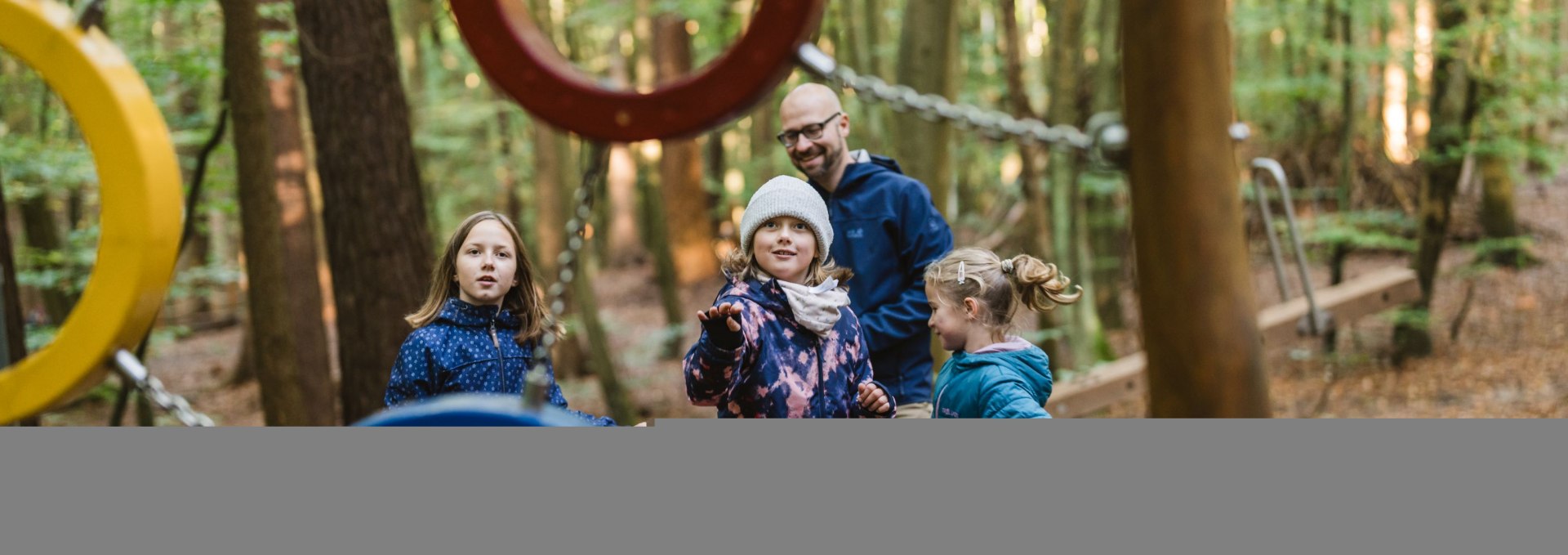 A group of children and an adult stand between colorful metal climbing rings on a forest playground.