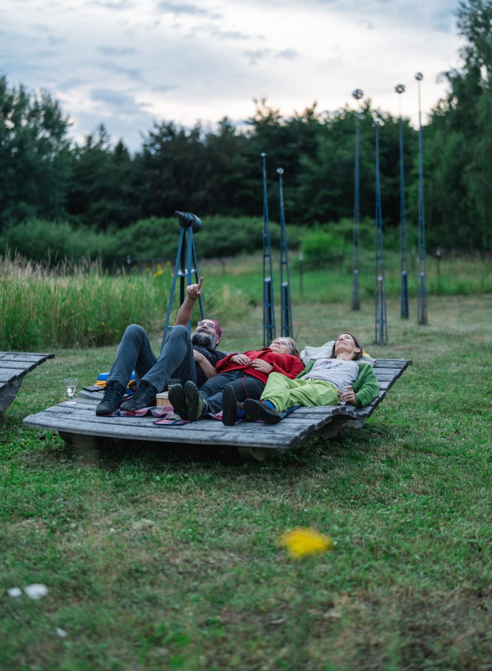 Trzy osoby leżą na drewnianej platformie pośród zieleni i spoglądają w wieczorne niebo – Park Gwiazd Meklemburgii-Pomorza Przedniego. // Na platformie widokowej trzy osoby leżą na trawie i podziwiają wieczorne niebo – w ciszy, z poczuciem przestrzeni, gotowe na zachwyt w Parku Gwiazd Meklemburgii-Pomorza Przedniego. // © MV-T/Gross Trzy osoby leżą na drewnianej platformie pośród zieleni i spoglądają w wieczorne niebo – Park Gwiazd Meklemburgii-Pomorza Przedniego.