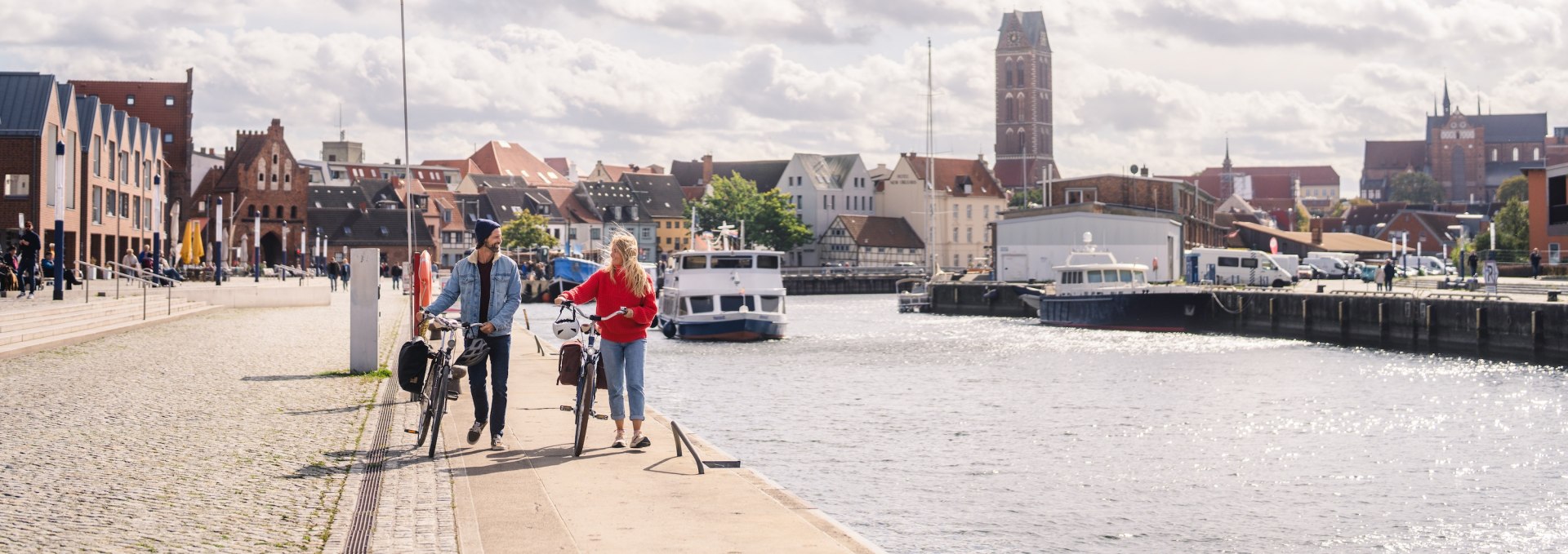 A couple push bicycles along Wismar's old harbor, with the city's historic skyline in the background.