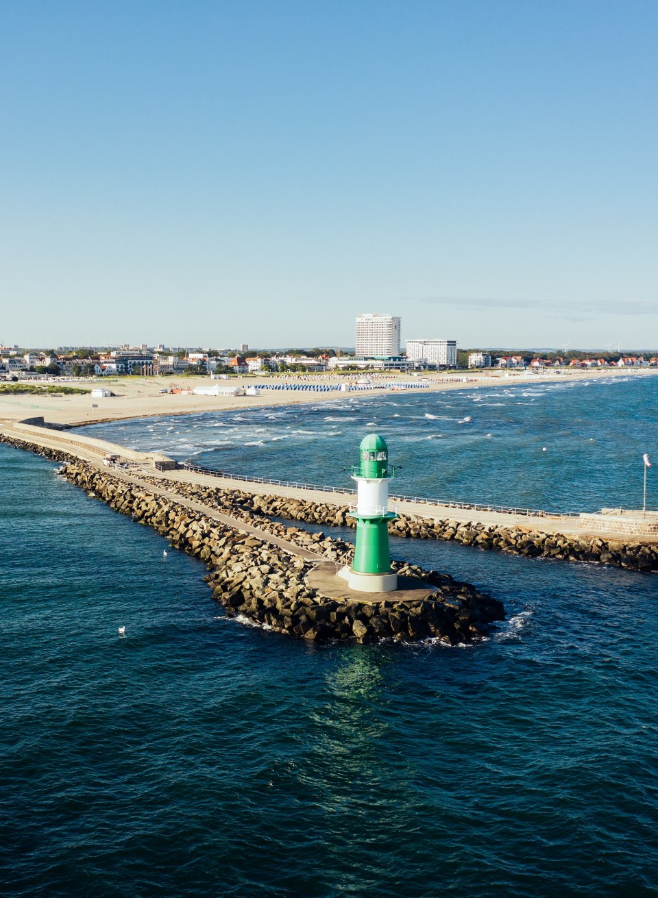 Green lighthouse on the pier of Warnemünde with a view of the beach, the Baltic Sea and the city panorama.