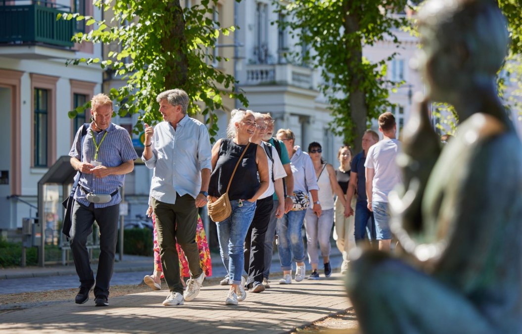 Een groep mensen loopt over een geplaveid pad met een stadsgids. Rechts, onscherp in beeld, is een standbeeld te zien bij de Pfaffenteich-vijver; de villa's verschijnen op de achtergrond. De focus ligt op de stadsgids en een geanimeerd pratend echtpaar., © Oliver Borchert