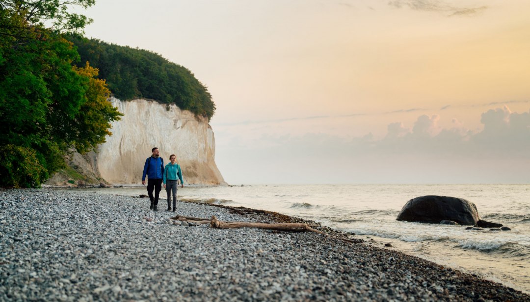 Frisse Oostzeelucht en schilderachtige uitzichten - een stel geniet van de rust tijdens een ochtendwandeling langs de krijtkust op het eiland Rügen. Perfect voor natuurliefhebbers en wandelliefhebbers., © TMV/Petermann Een paar wandelingen langs het stenen strand met uitzicht op de zee bij zonsopgang aan de krijtkust van Rügen.