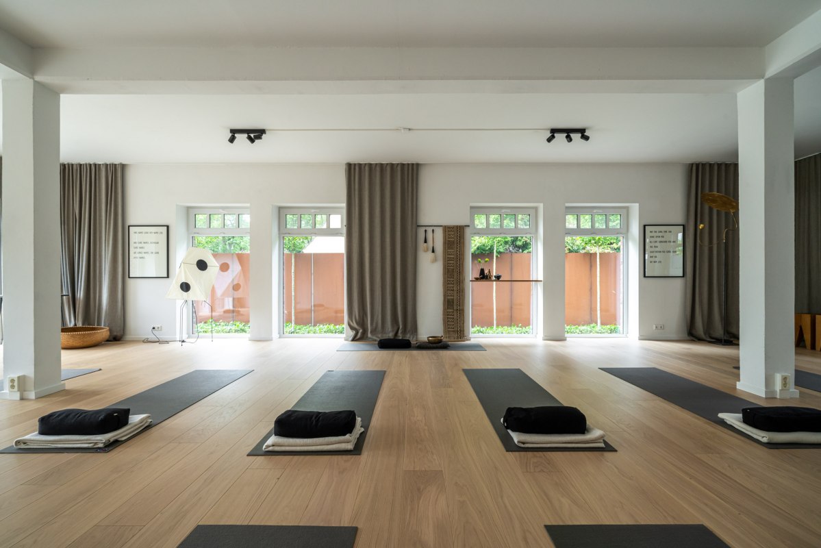 Een moderne, minimalistische yoga- en meditatieruimte met lichtgekleurde houten vloeren, grote ramen en subtiele decoraties. De matten liggen al klaar voor een komende yogasessie, vergezeld van zachte kussens en handdoeken., &copy; Hotel Carpe Diem