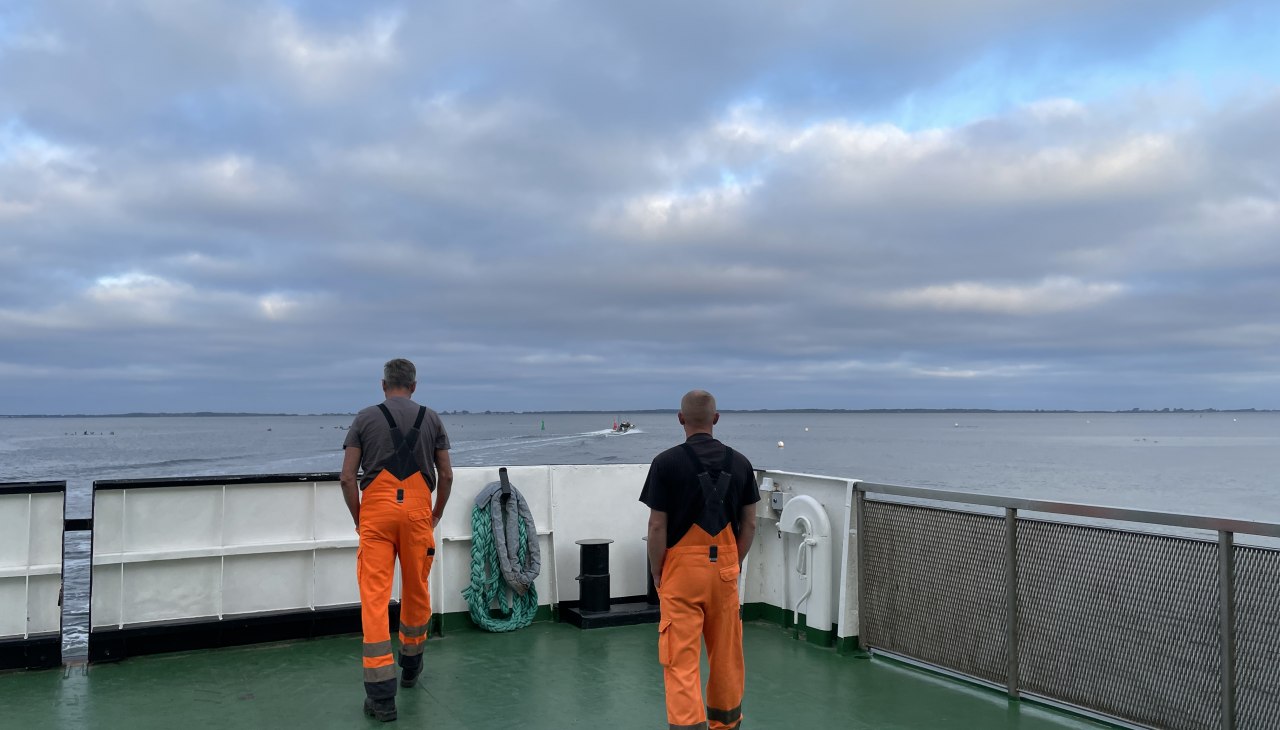 Op de veerboot staan twee arbeiders in oranje tuinbroeken bij de reling en kijken uit over het water., © Anja Heitmüller Op de veerboot staan twee arbeiders in oranje tuinbroeken bij de reling en kijken uit over het water., © Anja Heitmüller