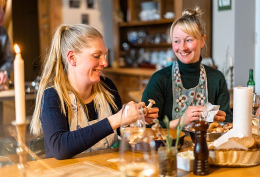 Twee vrouwen bereiden samen een maaltijd in de keuken van het landhuis Ehmkendorf, met kaarslicht en houten planken op de achtergrond. // Samen aan de lange houten tafel: verse ingrediënten gaan de pan in, gesprekken en kaarslicht vullen het landhuis Ehmkendorf. // © MV-T/Gross Twee vrouwen bereiden samen een maaltijd in de keuken van het landhuis Ehmkendorf, met kaarslicht en houten planken op de achtergrond.