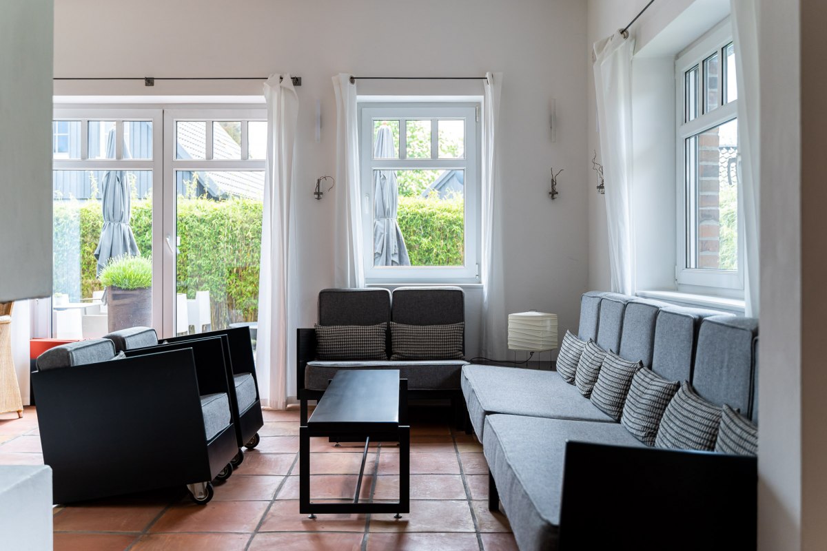 Een lichte en stijlvol ingerichte loungeruimte met grote ramen die veel natuurlijk licht binnenlaten. Grijze banken en fauteuils met minimalistische zwarte accenten nodigen je uit om te ontspannen. De warme terracotta vloeren en eenvoudige witte gordijnen geven de kamer een ontspannen en uitnodigende sfeer. // &copy; Hotel Carpe Diem