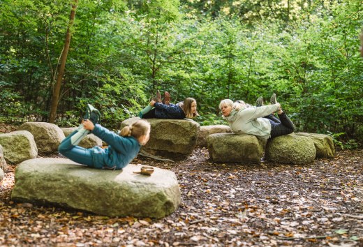 Kinderen en een volwassene liggen op grote stenen in het bos en doen samen yoga-oefeningen, omringd door dichte bomen en bladeren.