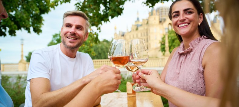 Gäste stoßen beim Winzerfest Schwerin auf dem Alten Garten mit einem Glas Wein an. Im Hintergrund erhebt sich das Schweriner Schloss und gibt dem Weinfest seine besondere Kulisse. // © LHSN/Celien Graubaum Gäste stoßen beim Winzerfest Schwerin auf dem Alten Garten mit einem Glas Wein an. Im Hintergrund erhebt sich das Schweriner Schloss und gibt dem Weinfest seine besondere Kulisse. // © LHSN/Celien Graubaum
