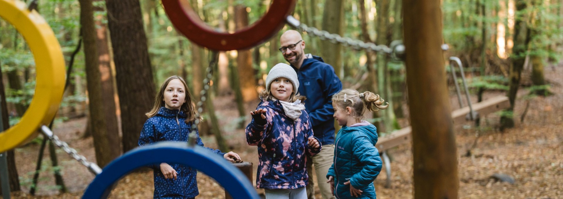 Een groep kinderen en een volwassene staan tussen kleurrijke metalen klimringen op een speelplaats in het bos.