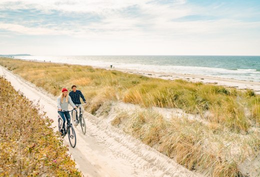 Two people cycle along a sandy dune path on the coast of Hiddensee, while waves crash on the beach and grass sways in the wind.