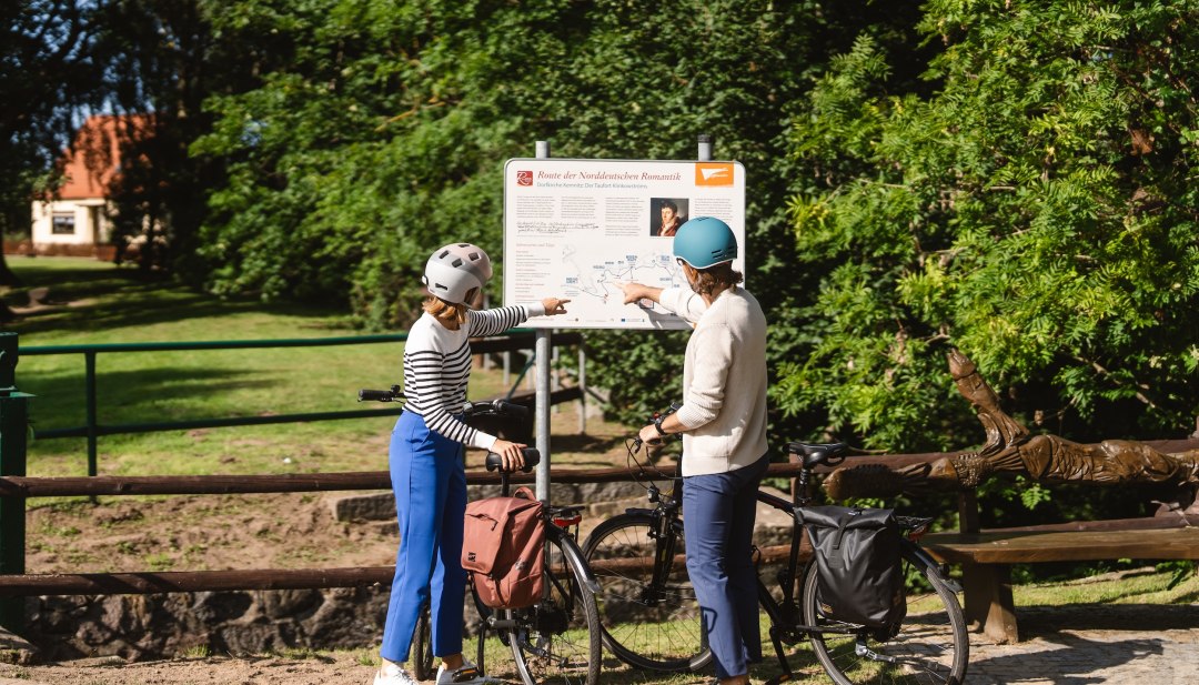 Dw&oacute;ch rowerzyst&oacute;w stoi przy znaku Szlaku P&oacute;łnocnoniemieckiego Romantyzmu