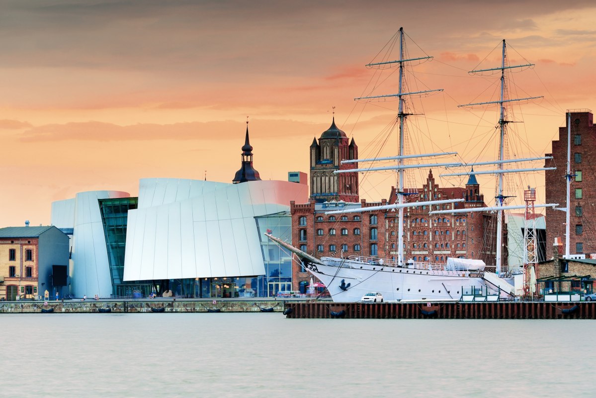 Nowoczesna architektura muzealna spotyka się z hanzeatyckim dziedzictwem: Ozeaneum w porcie Stralsund łączy się z historycznymi ceglanymi budynkami i żaglowcami, tworząc wyjątkową sylwetkę. W wieczornym świetle miasto wpisane na Listę Światowego Dziedzictwa UNESCO mieni się ciepłymi kolorami. Tutaj można poznać historię morską z bliska - od pokładu muzeum po promenadę portową. // &copy; Francesco Carovillano