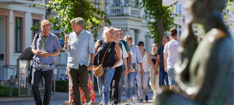 Een groep mensen loopt over een geplaveid pad met een stadsgids. Rechts, onscherp in beeld, is een standbeeld te zien bij de Pfaffenteich-vijver; de villa's verschijnen op de achtergrond. De focus ligt op de stadsgids en een geanimeerd pratend echtpaar., © Oliver Borchert