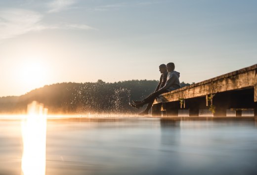 Twee mensen zitten bij zonsopgang op een steiger aan het Mirov-meer en dompelen hun voeten in het gladde water terwijl er een fijne mist over het oppervlak zweeft.