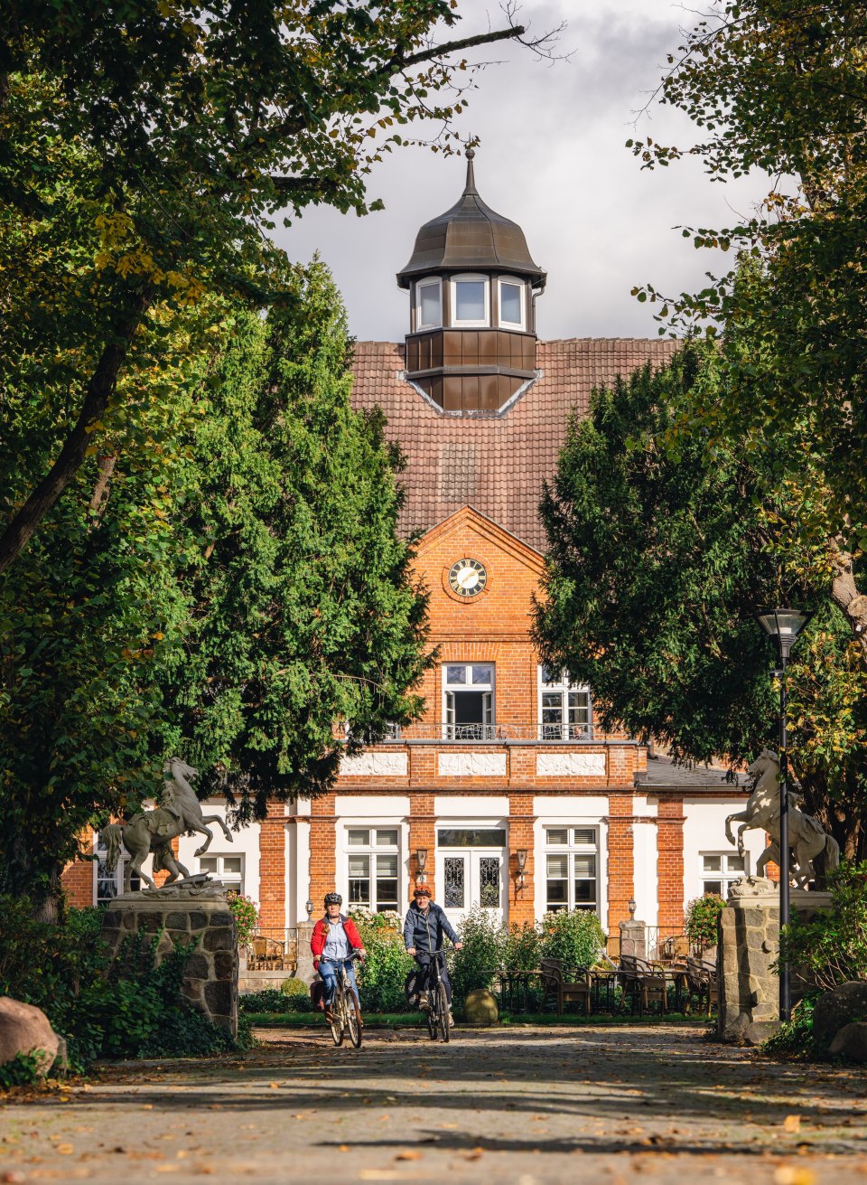 Twee fietsers naderen de ingang van Schloss Basthorst, omringd door bomen en een herfstsfeer.