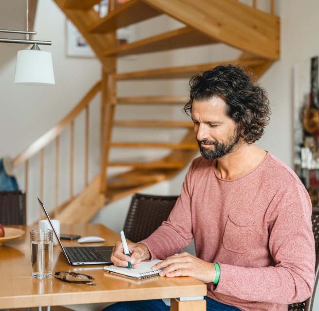A man sits at a wooden table in a vacation home, working on his laptop and taking notes - ideal conditions for a workation on the island of Poel. // Workation on the island of Poel: work productively in a cozy vacation home and enjoy the time out. // © MV-T/Gross A man sits at a wooden table in a vacation home, working on his laptop and taking notes - ideal conditions for a workation on the island of Poel.