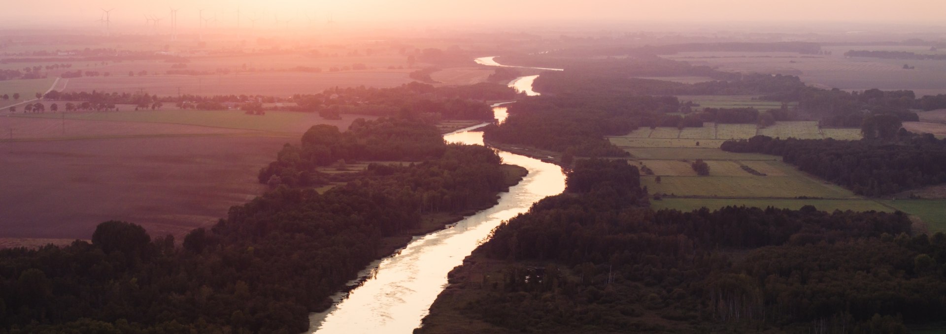Aerial view of the Peene at sunset, embedded in fields and forests, with an orange sky in the background. // Magical sunset - The Peene meanders through the idyllic landscape of Mecklenburg-Vorpommern. // © MV-T/Gross Aerial view of the Peene at sunset, embedded in fields and forests, with an orange sky in the background.