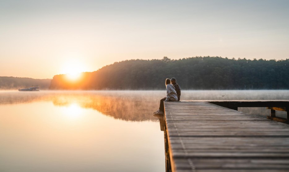 A couple sits on a jetty at sunset on Lake Mirov. A houseboat passes by in the background. // Sitting on the jetty enjoying the evening on Lake Mirower See // © MV-T/Gross A couple sits on a jetty at sunset on Lake Mirov. A houseboat passes by in the background.