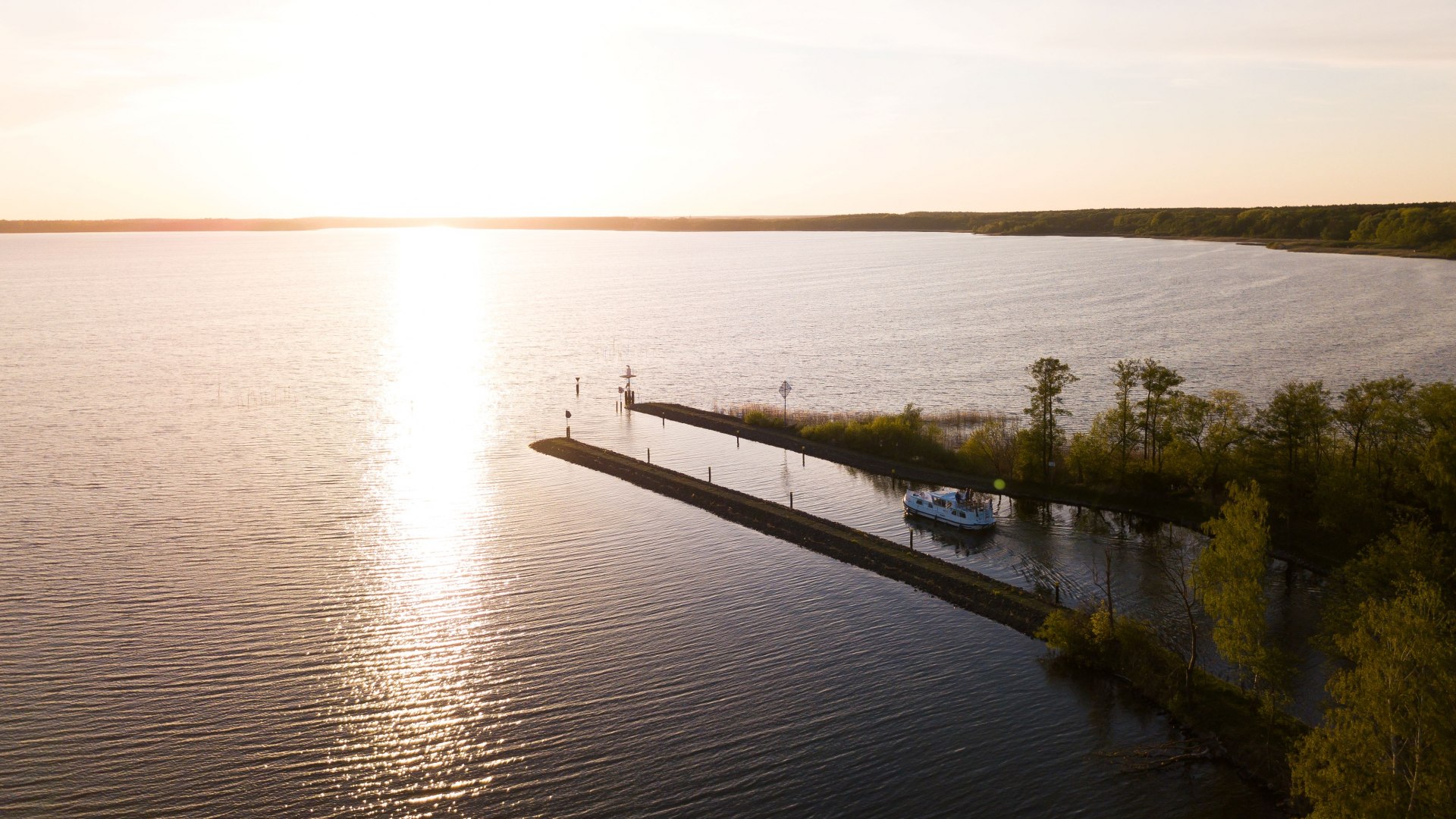 Luchtfoto van het Kölpin meer bij zonsondergang met een woonboot die door een smal kanaal vaart, omringd door bomen en rustig water.