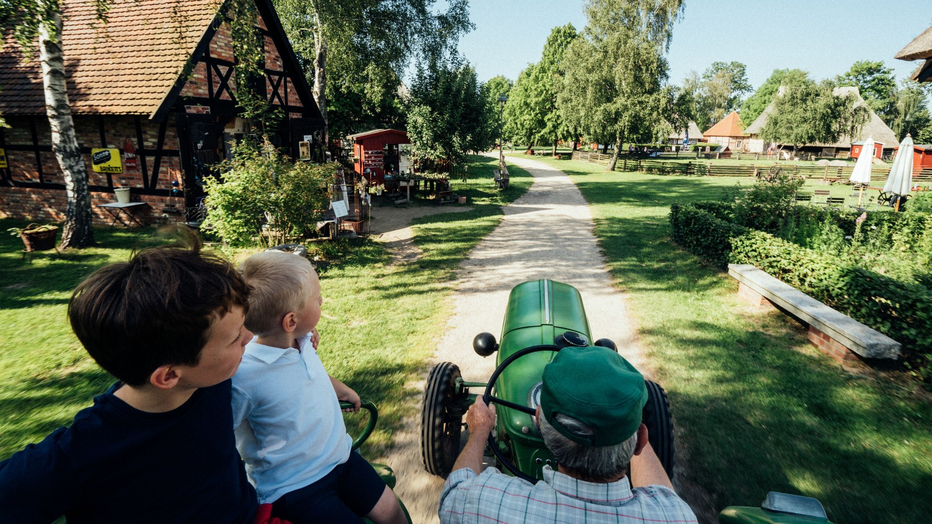 Dzieci jadą ze starszym mężczyzną na zabytkowym traktorze przez skansen Klockenhagen z domami z muru pruskiego i zielonym krajobrazem.