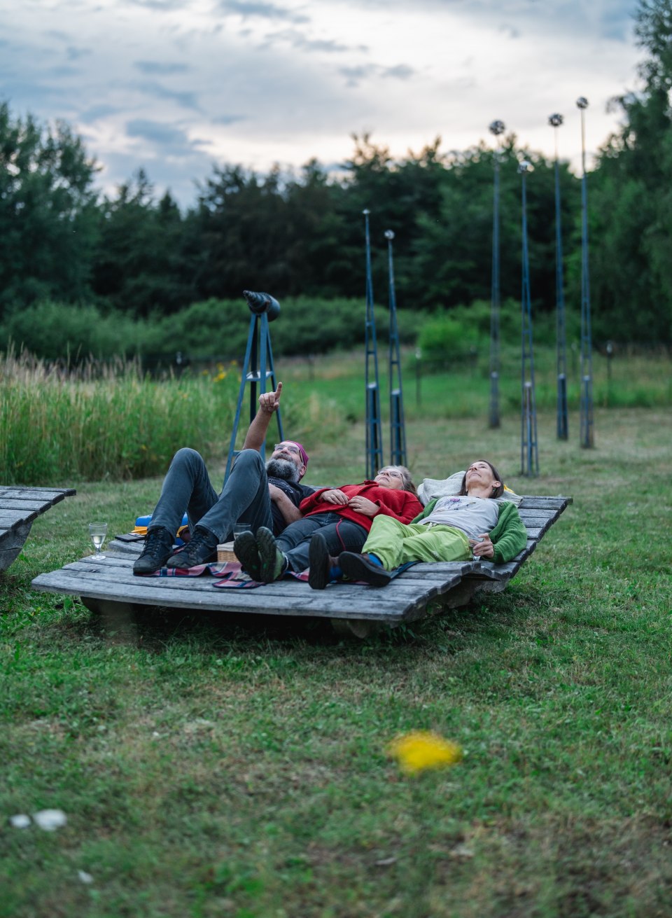 Tre personer ligger p&aring; en tr&auml;plattform mitt i naturen och blickar upp mot kv&auml;llshimlen, Stj&auml;rnparken Mecklenburg-Vorpommern.