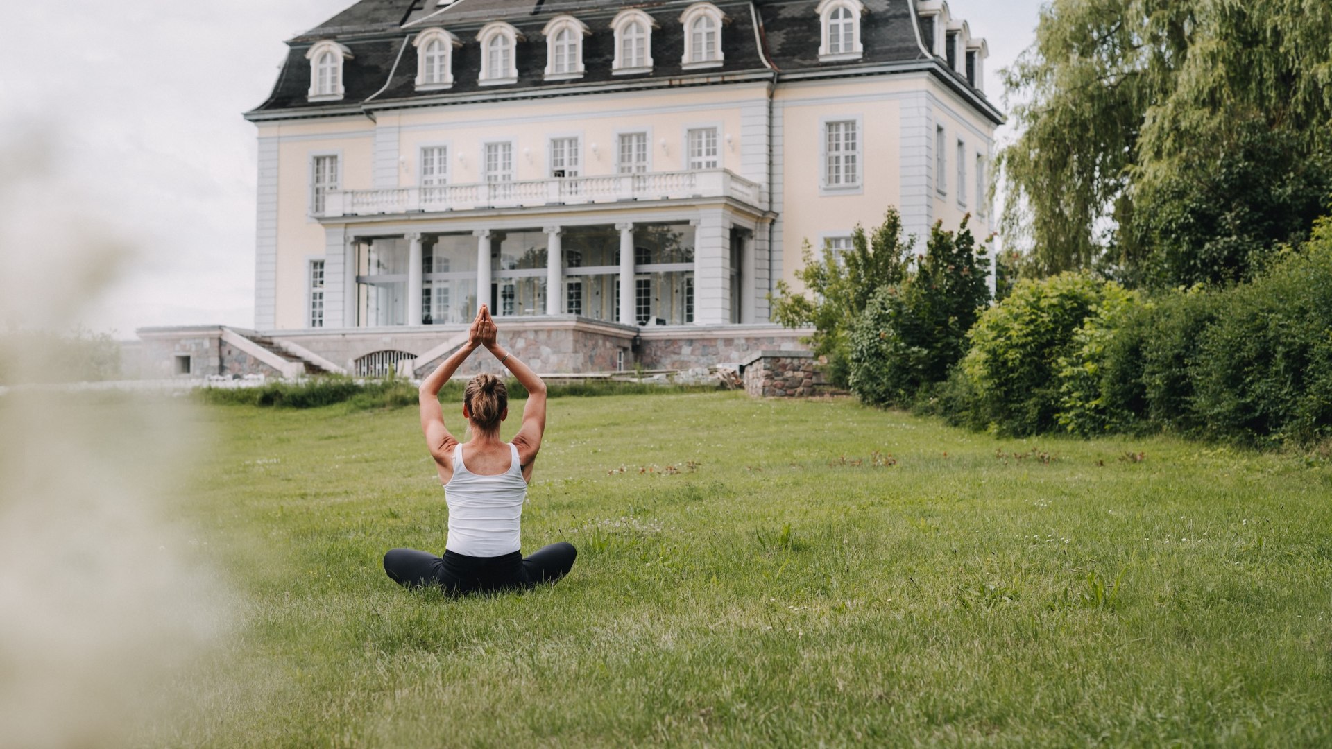 Woman meditating on a lawn in front of the historic mansion "The Reset Hotel" with white columns.