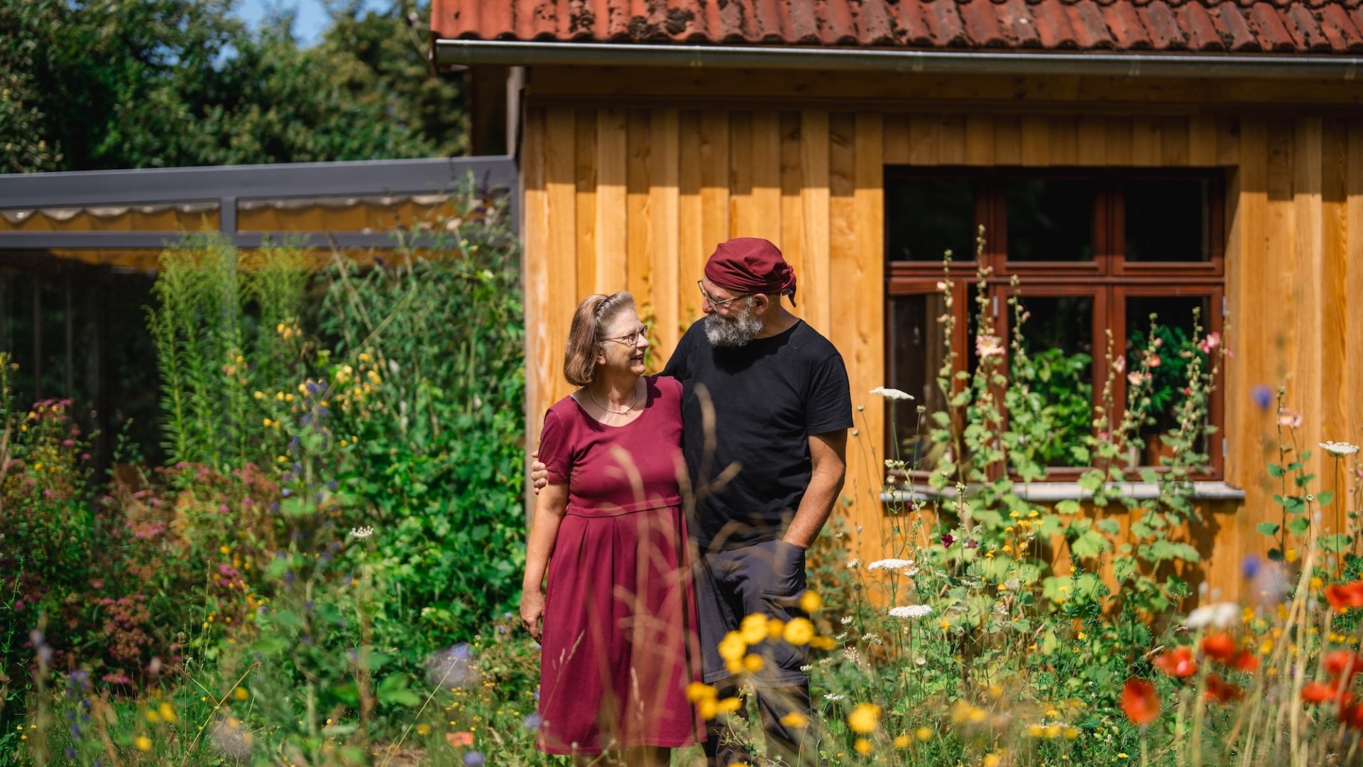 Et par står i en blomstrende have foran et træhus og smiler til hinanden, med farvestrålende vilde blomster i forgrunden. // Blandt vilde blomster og et træhus fanger blikket: Ralf Koch og hans kone står sammen i haven – nu er de ankommet til Mecklenburg-Vorpommern. // © MV-T/Gross Et par står i en blomstrende have foran et træhus og smiler til hinanden, med farvestrålende vilde blomster i forgrunden.