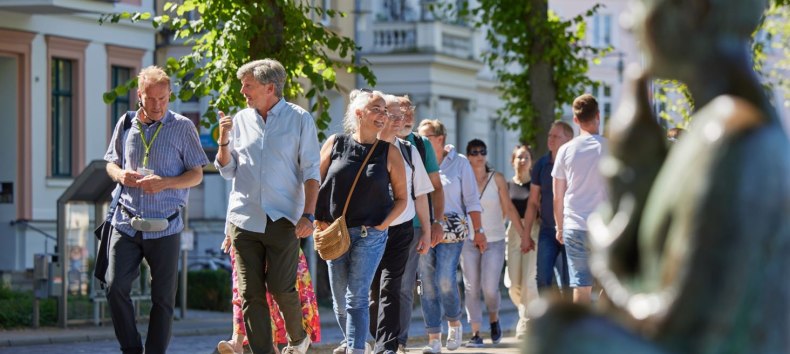 En gruppe mennesker går sammen med en byguide ad en brostensbelagt sti. Til højre, lidt ude af fokus, ses en statue ved Pfaffenteich, og i baggrunden dukker villaerne op. Fokus er rettet mod byguiden og et par, der fører en livlig samtale. // © Oliver Borchert En gruppe mennesker går sammen med en byguide ad en brostensbelagt sti. Til højre, lidt ude af fokus, ses en statue ved Pfaffenteich, og i baggrunden dukker villaerne op. Fokus er rettet mod byguiden og et par, der fører en livlig samtale. // © Oliver Borchert