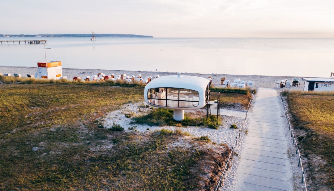 Luchtfoto van de Müther Tower op het strand van Binz met de Baltische Zee en strandstoelen. // De Müther Tower troont direct op het strand in Binz - een uniek stukje architectuur tussen de duinen en de Baltische Zee. De futuristische vorm steekt af tegen het zachte zand, terwijl strandstoelen langs de kust staan. Een plek die moderniteit en uitzicht op zee combineert. // © MV-T/Ulrich Luchtfoto van de Müther Tower op het strand van Binz met de Baltische Zee en strandstoelen.