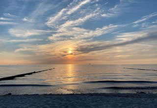 The picture shows the mirror-smooth Baltic Sea, whose calm water surface reflects the colors of the sunset. The sky is partly light blue, interspersed with fine streaky clouds that lend the panorama a gentle dynamism. A peaceful beach stretches along the edge of the motif, inviting the viewer to linger. The atmosphere is calm and harmonious, as if the world is pausing for a moment to enjoy the beauty of the moment. // &copy; byc