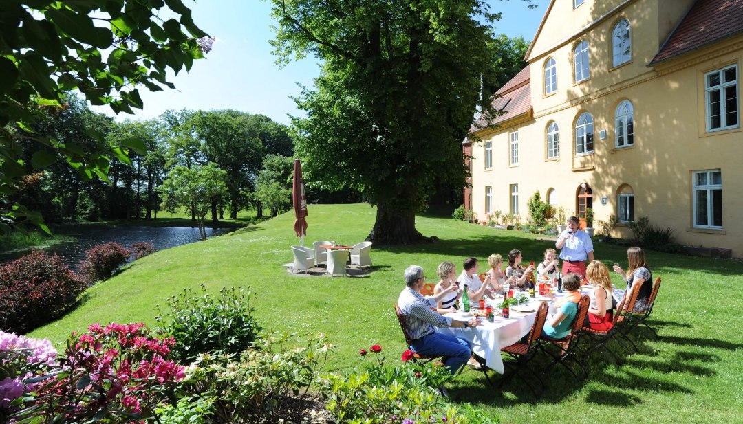 Mensen zitten in een tuin voor een landhuis met een lange tafel en picknicken in de zon. // Culinaire hoogstandjes en stimulerende gesprekken op kasteel Lühburg // © MV-T/Foto@Andreas-Duerst.de Mensen zitten in een tuin voor een landhuis met een lange tafel en picknicken in de zon.