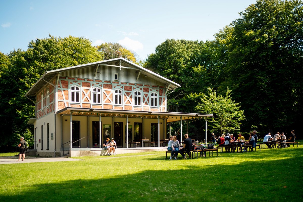 Een lichtgekleurd gebouw met een terras, waarvoor verschillende mensen op bankjes op een groen gazon zitten. // © Nationalpark-Zentrum KÖNIGSSTUHL / Dan Petermann Een lichtgekleurd gebouw met een terras, waarvoor verschillende mensen op bankjes op een groen gazon zitten. // © Nationalpark-Zentrum KÖNIGSSTUHL / Dan Petermann