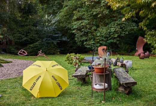 Sculpture garden with yellow KunstOffen umbrella and metal sculptures at Kunst-Werk Kummer. In front of it is a yellow umbrella with the inscription "KunstOffen"