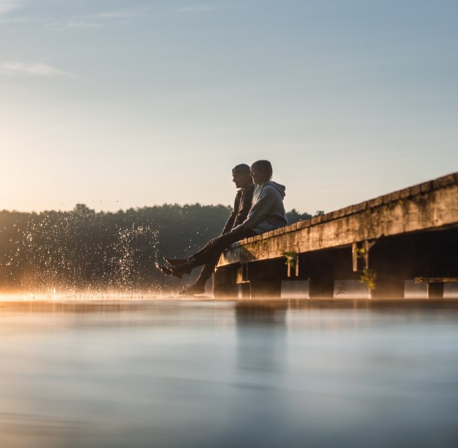 Two people sit on a jetty on Lake Mirov at sunrise and dip their feet into the smooth water while a fine mist hovers over the surface.