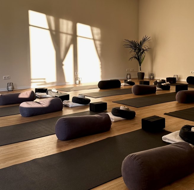 Bright, minimalist yoga room with wooden flooring, several yoga mats, meditation cushions, bolsters and yoga blocks. Warm sunlight shines through large windows and creates a calm, relaxing atmosphere for yoga, meditation and relaxation. // &copy; MOVA PLACE - Yoga & Co. Studio