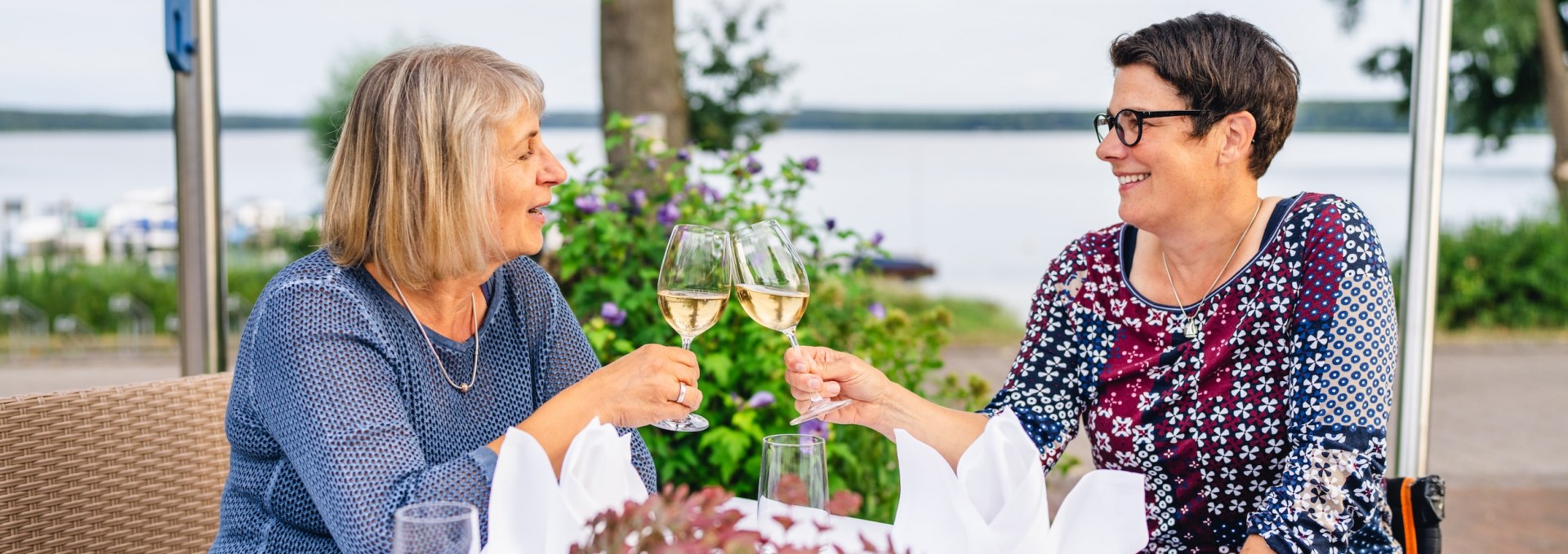 Przy stole, przy kt&oacute;rym siedzą dwie kobiety, wznoszony jest toast kieliszkiem szampana.