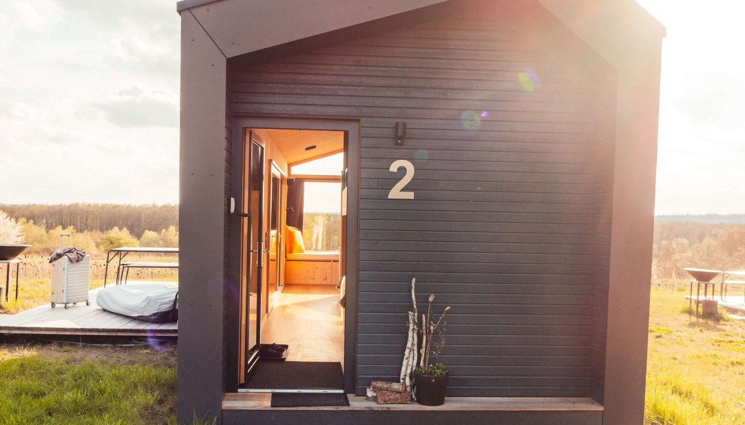 Stylowy minimalizm spotyka się z naturą z zewnątrz o zachodzie słońca - Tiny House z panoramicznymi oknami na relaksującą przerwę., © mari.linni || Nicola Link Berlin Mały, ciemny domek z drewnianą boazerią, otwartymi drzwiami wejściowymi i promieniami słońca na zielonej łące.