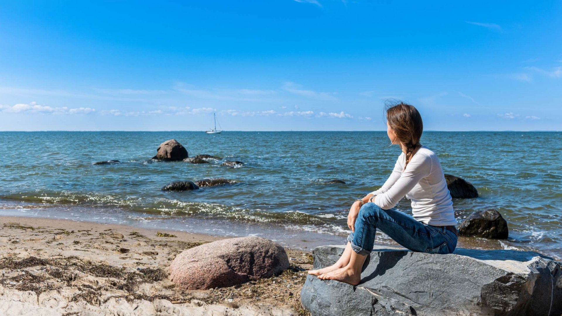 Een model voor een schets: Uitzicht op de Oostzee vanaf het strand. Er liggen een handvol grote stenen op het strand en aan de horizon ligt een zeilboot.