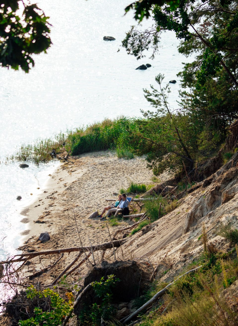 A small sandy beach in the Southeast Rügen Biosphere Reserve, surrounded by steep banks and dense vegetation. Two people sit relaxed on a bench with a view of the glistening water interspersed with scattered rocks. A quiet and secluded atmosphere invites you to linger.