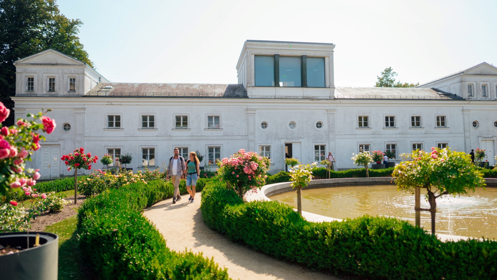 Een echtpaar wandelt over een goed onderhouden pad door de tuin van de oranjerie in Putbus. De tuin is versierd met rozen en buxushagen, terwijl op de voorgrond een kleine vijver met een fontein te zien is. Op de achtergrond staat het historische oranjeriegebouw met zijn witte gevels en grote ramen.