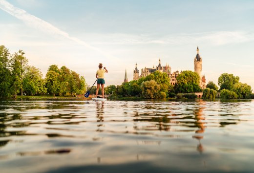 Een persoon peddelt op een meer in het warme avondlicht en kijkt uit over een historisch kasteel omringd door weelderig groen. // Glijden over het meer van Schwerin creëert een stil, krachtig moment waarin de natuur en de achtergrond van het kasteel samensmelten. // © MV-T/Petermann Een persoon peddelt op een meer in het warme avondlicht en kijkt uit over een historisch kasteel omringd door weelderig groen.